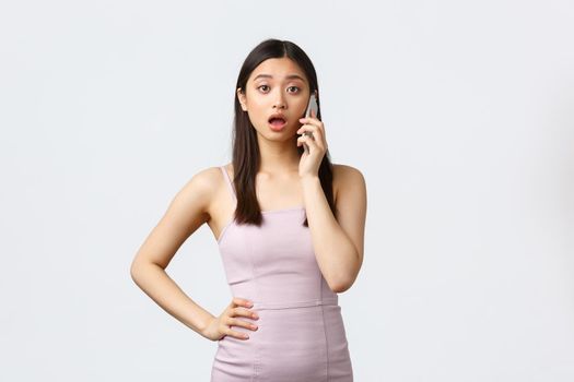 Luxury women, party and holidays concept. Surprised and shocked asian woman in dress having phone conversation, calling someone and gossiping, standing white background