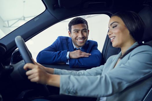 So, what do you think. an attractive young woman sitting in a new car while speaking to a handsome male car salesman about a deal.