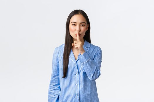 Smiling cunning cute asian girl in blue pajamas arrange secret surprise for sleepover girlfriends, showing shush gesture, hushing want silence, asking be quiet, standing over white background