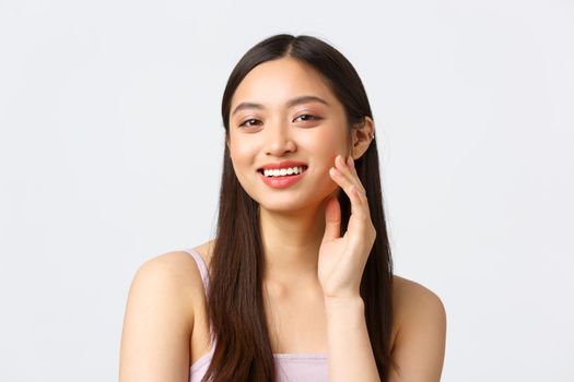 Beauty, fashion and people emotions concept. Beautiful asian girl trying new skincare routine product or makeup, smiling happy and satisfied as touching skin gently, white background