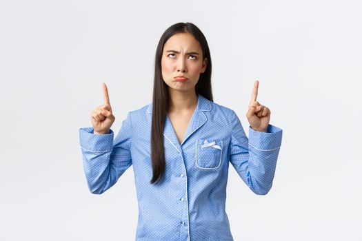 Complaining and bothered asian girl pouting and frowning displeased, looking and pointing fingers up disappointed, standing in blue pajamas, standing unsatisfied white background
