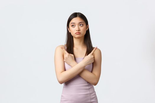 Luxury women, party and holidays concept. Indecisive pretty asian girl in evening dress making tough decision, pointing fingers sideways at both variants, being confused, white background