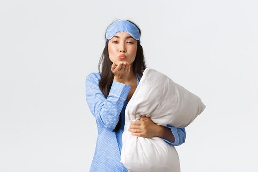 Smiling romantic asian girl in blue pajama and sleeping mask, hugging pillow and sending goodnight air kiss at camera with coquettish, sensual face, standing white background