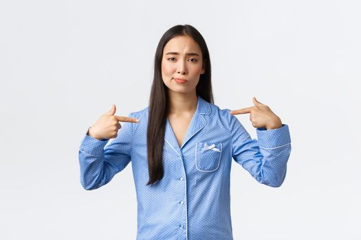Indecisive and unconfident asian girl in blue pajamas smirk uncertain and pointing at herself, being unsure own abilities, talking about herself, standing white background