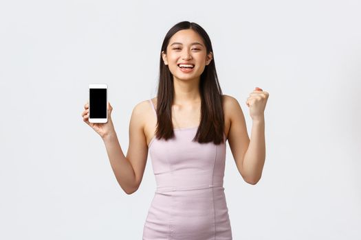 Luxury women, party and holidays concept. Rejoicing, happy asian girl in dress, feeling happy and delighted, got special offer notification on smartphone screen, chanting and show mobile phone
