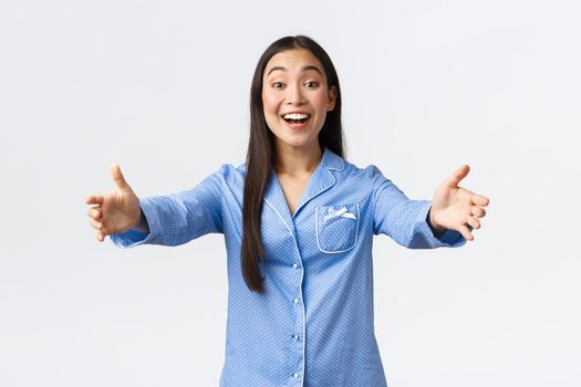 Friendly asian girl in blue pajamas welcome girlfriends to her sleepover party, smiling happy and reaching hands forward to greet or hug person, standing in pajamas over white background