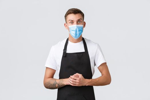 Coronavirus, social distancing in cafes and restaurants, business during pandemic concept. Serious handsome barista in medical mask, salesman listening to customer over counter, white background