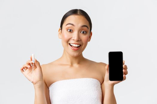 Beauty, personal and intimate care, mobile application concept. Cheerful smiling asian woman showing her menstrual cycle, period tracker on smartphone screen, holding tampon