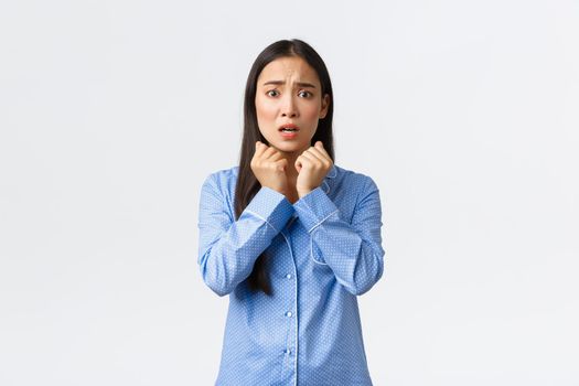 Scared timid asian girl panicking, looking anxious and afraid, shivering as standing in blue pajamas, hear strange noise being home alone, standing over white background alarmed