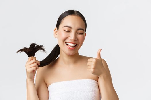 Beauty, hair loss products, shampoo and hair care concept. Close-up of satisfied, happy asian girl in bath towel showing thumbs-up and healthy hair ends, standing pleased white background