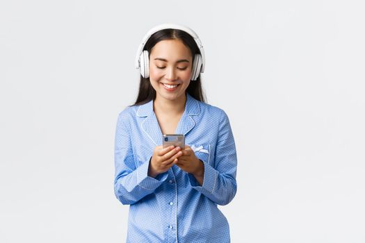 Home leisure, weekends and lifestyle concept. Smiling pretty asian woman in blue pajama and headphones, listening music, texting or watching video on mobile phone, smile at display