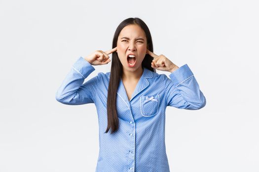 Angry and pissed-off young asian woman in blue pajamas screaming with ears shut, plug fingers from disturbing loud noise at night, complaining neighbour music, cant sleep, standing white background