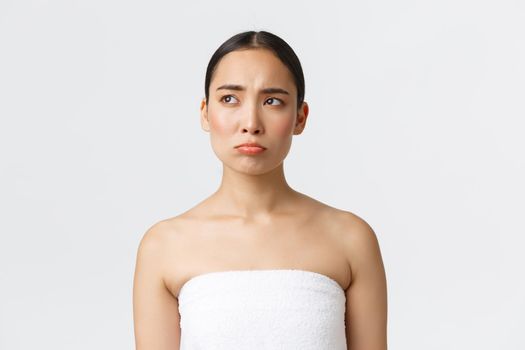 Beauty, cosmetology and spa salon concept. Pouting sad and disappointed asian girl in white towel sulking and looking left with regret, see something upsetting, being jealous