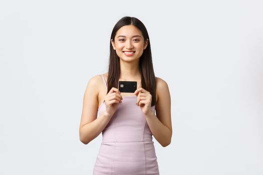 Luxury women, party and holidays concept. Happy rich pretty asian girl in evening dress, showing her brand new credit card, smiling pleased as going shopping, paying for order contactless
