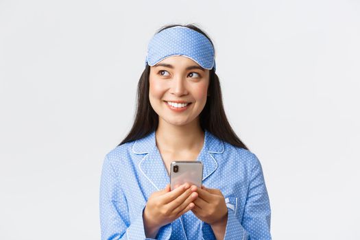Close-up of beautiful asian girl in sleeping mask and blue pajama getting ready sleep, looking away thoughtful as using mobile phone, messaging or using social network to write post, white background