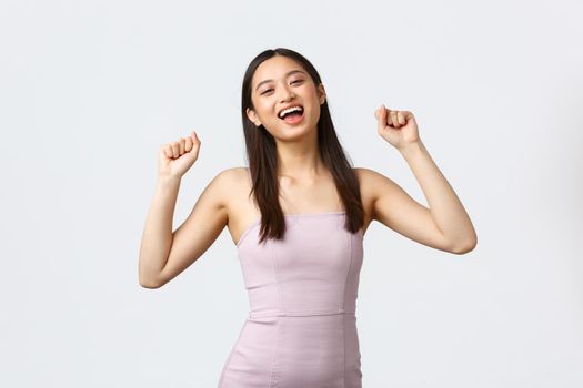 Luxury women, party and holidays concept. Relieved and happy pretty korean girl in evening dress, raise hands up shouting yes delighted, winning, become champion, triumphing over white background