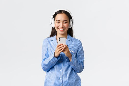 Home leisure, weekends and lifestyle concept. Smiling pretty asian girl in blue pajama listening music wireless headphones and taking photo on mobile phone, standing white background