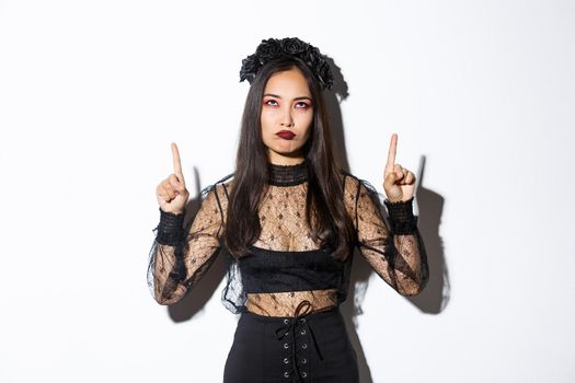 Grumpy asian female in witch costume looking disappointed and upset, pointing fingers up and sulking displeased, standing over white background in halloween costume