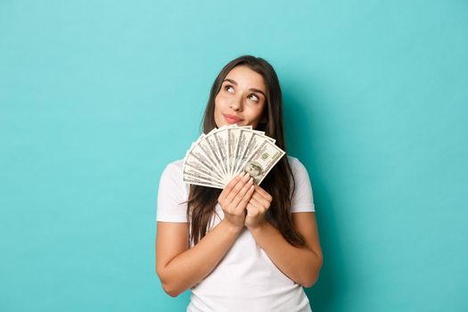 Portrait of attractive brunette woman in white t-shirt, dreaming about shopping, holding money, standing over blue background
