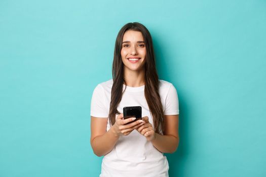 Portrait of attractive smiling woman in white t-shirt, holding smartphone and looking at camera, standing over blue background