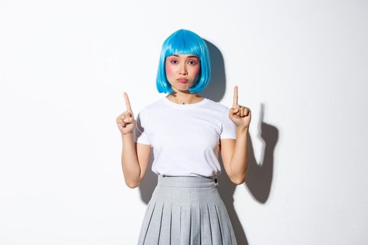 Disappointed gloomy asian girl in blue wig pointing fingers up, sulking upset and showing something bad, negative emotion, standing over white background