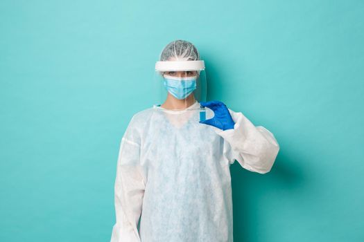 Concept of coronavirus, healthcare and quarantine. Image of female doctor in personal protective equipment, showing covid-19 vaccine, standing over blue background
