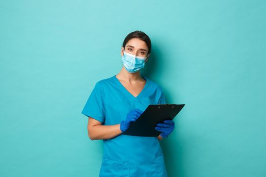 Covid-19, pandemic and medicine concept. Image of beautiful female doctor in medical mask, gloves and scrubs, provide patient check up, writing down something on clipboard, blue background