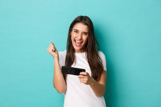 Image of happy young woman in white t-shirt, playing mobile phone game and winning, rejoicing while standing over blue background