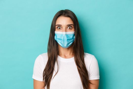 Concept of coronavirus, health and social distancing. Image of shocked young woman in medical mask, looking amazed at camera, standing over blue background