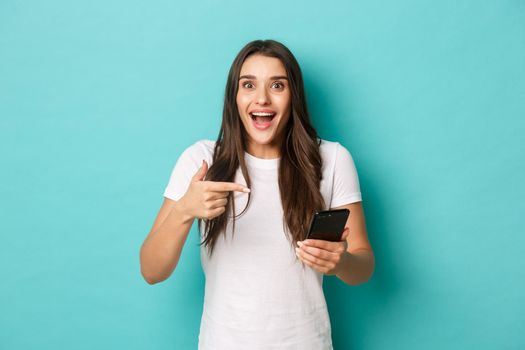 Portrait of excited attractive woman with long dark hair, wearing white t-shirt, pointing finger at mobile phone, showing awesome promo, standing over blue background