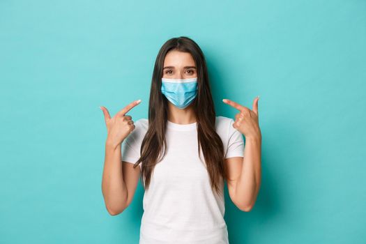 Concept of pandemic, covid-19 and social distancing. Smiling friendly girl in white t-shirt, recommend to wear medical mask during coronavirus, standing over blue background