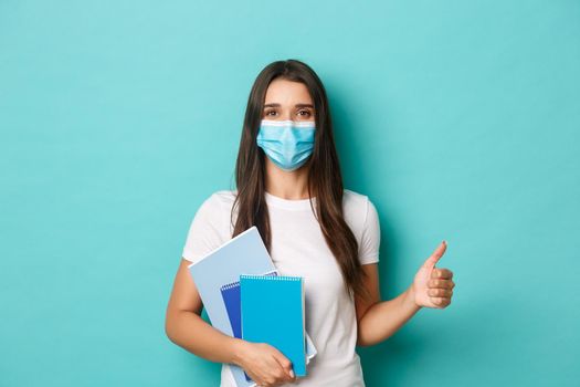 Concept of coronavirus, health and social distancing. Portrait of female student attend classes in medical mask, showing thumbs-up in approval, holding notebooks, standing over blue background