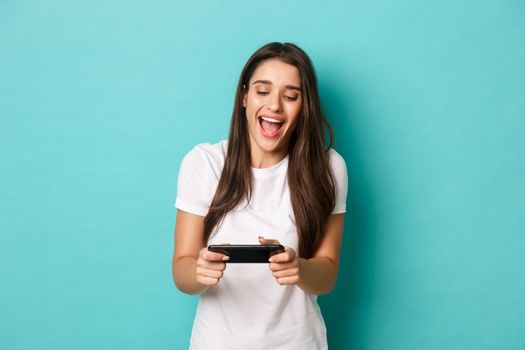 Image of happy young woman in white t-shirt, playing mobile phone game and smiling, standing over blue background