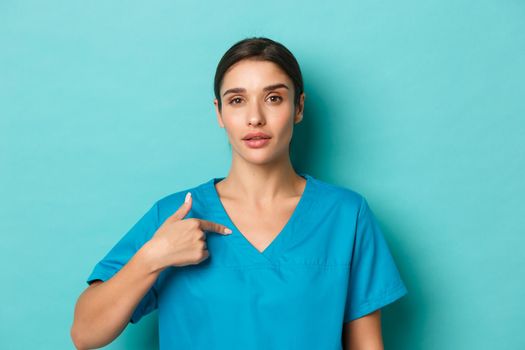Coronavirus, social distancing and health concept. Close-up of confident female doctor in scrubs, pointing at herself, working during pandemic, standing over blue background