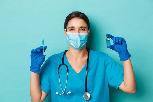 Concept of covid-19 and quarantine concept. Cheerful smiling female doctor in medical mask, latex gloves and scrubs, showing syringe filled with coronavirus vaccine, standing over blue background
