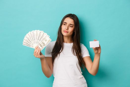Image of thoughtful young woman making choice between cash and credit card, standing over blue background