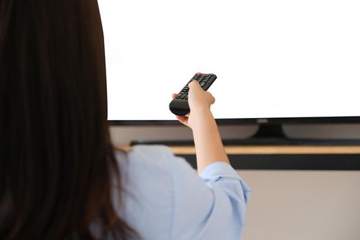Young woman holding a remote to turn on the TV in the New Normal era where she can't go out of the house, the white space on the TV can be inserted into text or images.