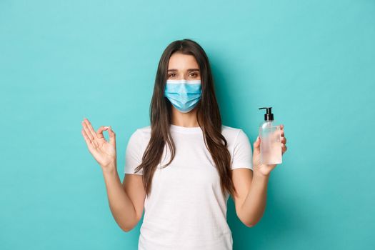 Concept of coronavirus, health and social distancing. Image of young brunette woman in medical mask, recommending to use hand sanitizer or antiseptic, showing okay sign