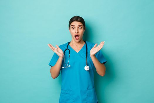 Coronavirus, pandemic and social distancing concept. Image of surprised beautiful female doctor in scrubs, open mouth amazed and looking at something, standing over blue background