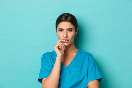 Coronavirus, social distancing and health concept. Close-up of serious thoughtful female doctor, wearing scrubs, frowning and looking at camera, thinking or making decision, blue background