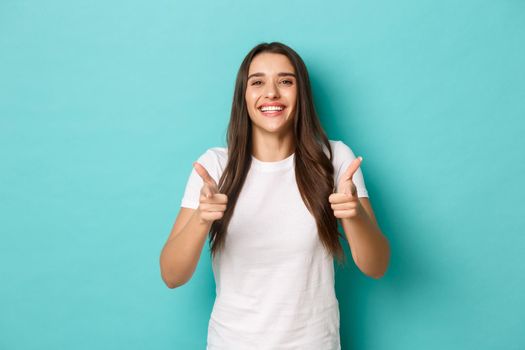 Cheerful brunette woman in white t-shirt, congratulating you, pointing fingers at camera, smiling pleased, praising good job, standing over blue background