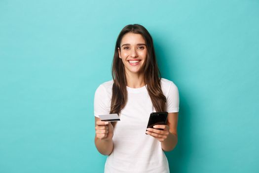 Portrait of young brunette woman in casual white t-shirt, shopping online, holding credit card and mobile phone, standing over blue background