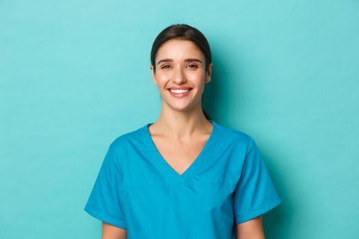 Coronavirus, social distancing and health concept. Close-up of young smiling female doctor, wearing scrubs, looking cheerful, standing over blue background
