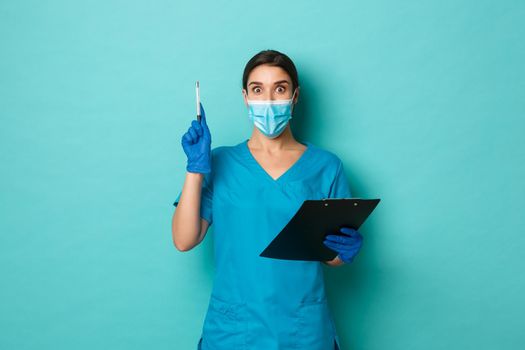 Covid-19, pandemic and medicine concept. Image of beautiful female doctor in medical mask, gloves and scrubs, holding clipboard and raising pen in eureka sign, having an idea or solution