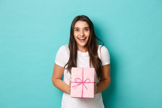 Concept of holidays and celebration. Image of excited woman looking happy, smiling and receiving gift wrapped in pink box, standing over blue background