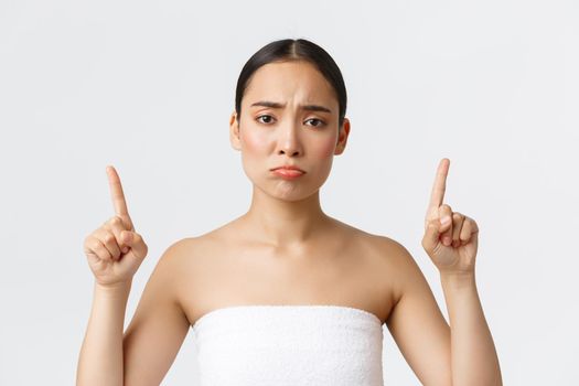 Beauty, cosmetology and spa salon concept. Disappointed pouting asian girl in white towel complaining or being jealous, pointing fingers up and whining about promo, white background