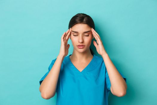 Coronavirus, social distancing and health concept. Close-up of woman doctor in scrubs feeling exhausted, having a headache, standing over blue background