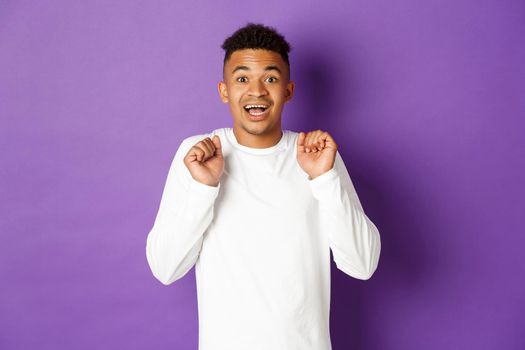 Image of hopeful african-american man, looking with anticipation at camera, smiling and jumping from excitement, standing over purple background