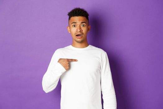 Portrait of confused and nervous african-american young man, pointing at himself, being chosen, standing over purple background