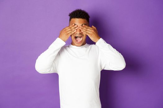 Image of handsome african-american 20s guy, cover eyes with hands and smiling, waiting for surprise with excited face, standing over purple background